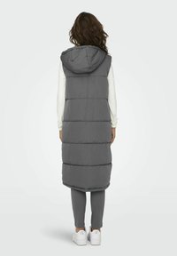 Gilet rembourré gris avec capuche, présentant un design long et matelassé ainsi qu'une texture lisse. Porté par-dessus une chemise à manches longues de couleur claire et des leggings gris.