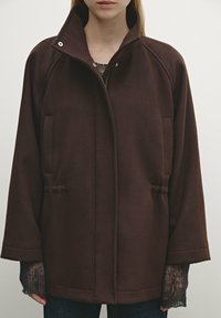 Donna che indossa una giacca zip-up larga di colore marrone scuro con colletto alto e maniche lunghe sopra un maglione grigio a trama e pantaloni scuri.