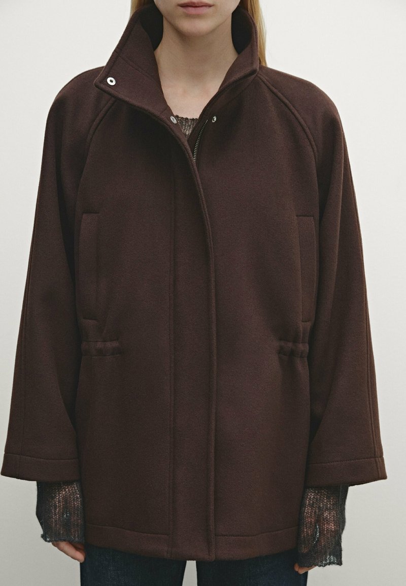 Donna che indossa una giacca zip-up larga di colore marrone scuro con colletto alto e maniche lunghe sopra un maglione grigio a trama e pantaloni scuri.