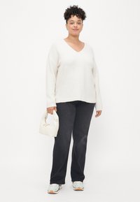 Licht beige V-hals trui, ontspannen pasvorm, gecombineerd met donkerblauwe jeans. Model houdt een kleine witte handtas vast. Sneakers met turquoise accenten aan de voeten.