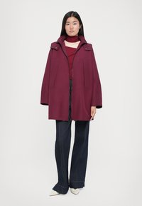 Manteau long bordeaux avec capuche, doté de deux poches avant et d'une coupe décontractée, assorti à un jean foncé évasé et des chaussures blanches à bout pointu.