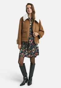 Brauner Wollmantel mit weißem Kragen, ausgestattet mit einem Reißverschluss und einer Fronttasche, getragen über einem marineblauen Blumenkleid. Schwarze kniehohe Stiefel komplettieren den Look.
