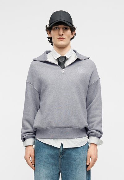 Νεαρός άνδρας που φοράει μαύρο καπέλο, γκρι φούτερ με φερμουάρ έως το στήθος πάνω από ένα πουκάμισο με γιακά και μαύρη γραβάτα, και μπλε τζιν, στέκεται μπροστά σε απλό φόντο.