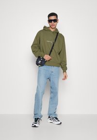 Oliven grønn hettegenser med logo, lyseblå jeans, svart og hvit sneakers, og en liten svart veske. Modellen har på seg solbriller og står vendt mot kameraet.