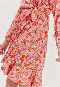 Kvetinové šaty v ružových a oranžových odtieňoch, s viazaním v páse, dlhými rukávmi a volánovým lemom. Materiál vyzerá ľahký a s potlačou.