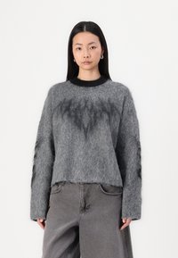 Grijze gebreide trui met een fuzzy textuur, voorzien van een zwarte ribbelkraag en donkergrijze accenten. Combinatie met grijze losse broeken.