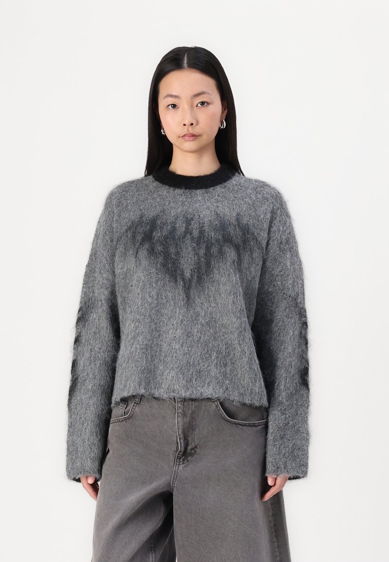 Grijze gebreide trui met een fuzzy textuur, voorzien van een zwarte ribbelkraag en donkergrijze accenten. Combinatie met grijze losse broeken.