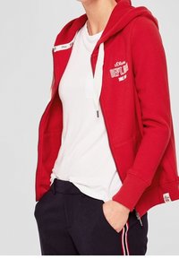 Rød zip-up hættetrøje med en frontlomme, drawstring hætte og hvidt logo print på brystet; parret med marineblå bukser med sidelommer.