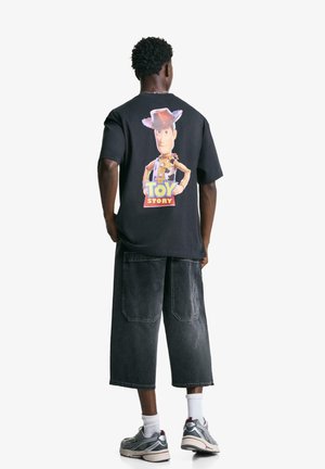 Tricou negru din bumbac cu un grafic mare cu Woody din Povestea Jucăriilor și textul "POVESTEA JUCĂRIILOR" în partea de jos, purtat cu pantaloni scurți din denim gri.
