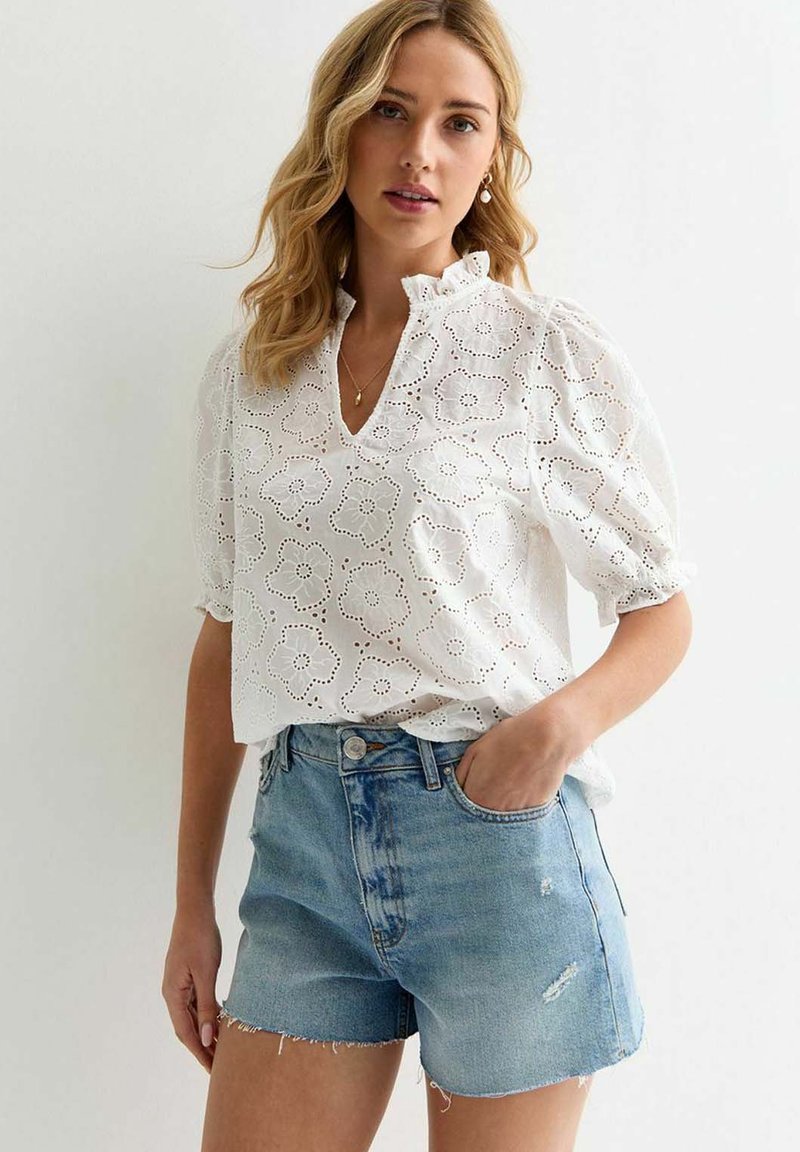 Witte katoenen blouse met een bloemenooglettend patroon, korte pofmouwen en een V-hals, gecombineerd met lichtblauwe versleten denim shorts.