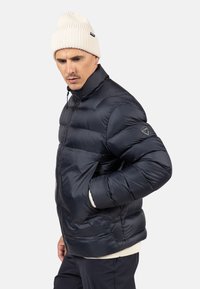 Giacca imbottita blu navy con colletto alto e polsini elasticizzati, con una toppa con logo sulla manica e chiusura frontale con zip. Indossata con un berretto color crema.