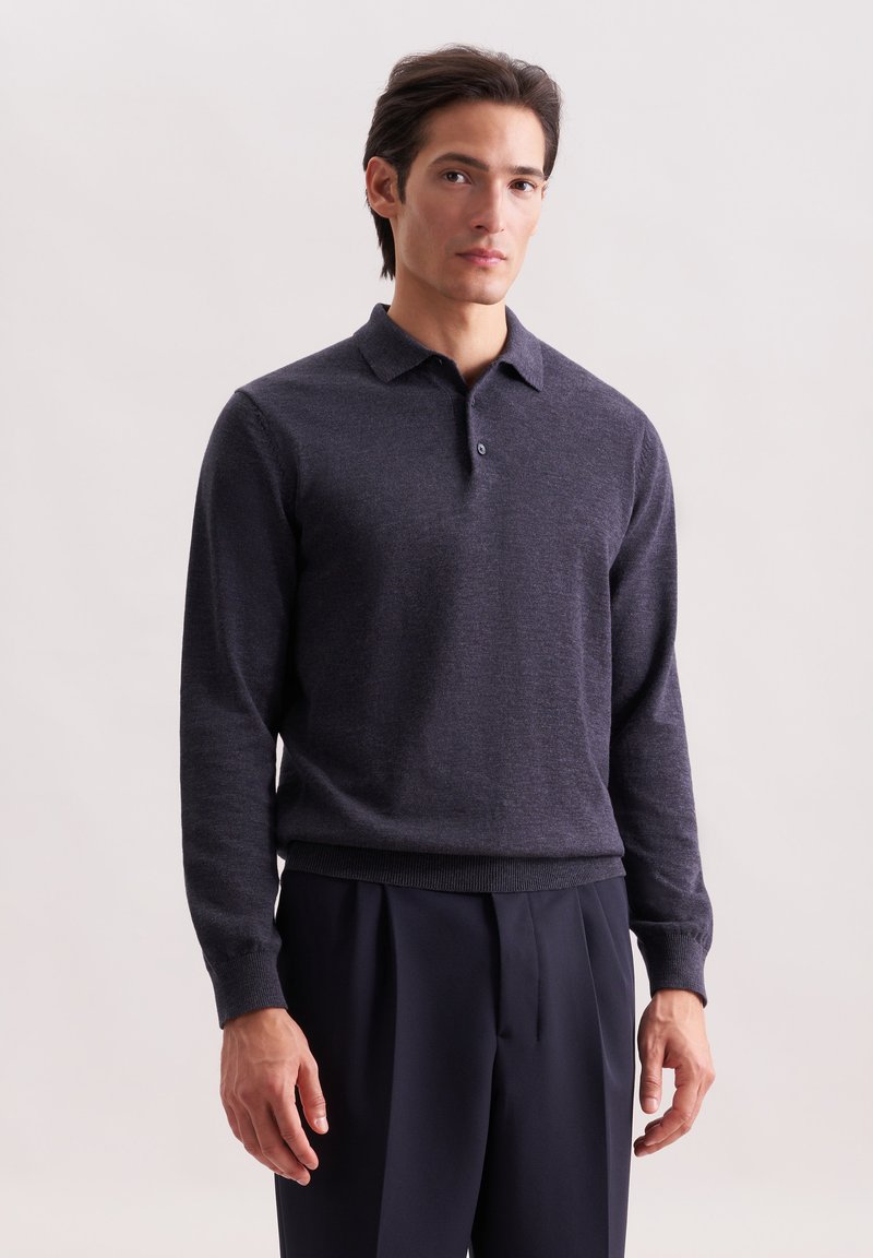 Homme portant un pull polo à manches longues gris foncé et un pantalon noir plissé, se tenant contre un fond clair uni.