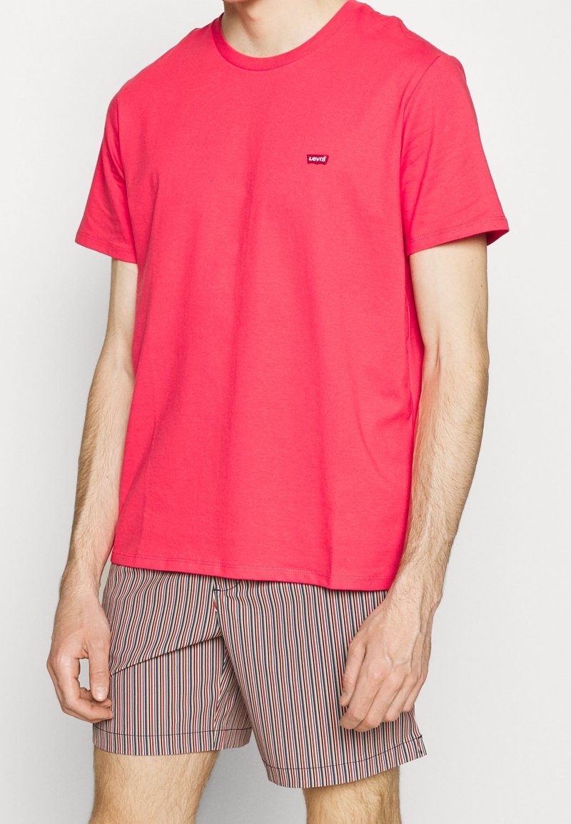 Homme portant un t-shirt Levi's rouge vif et un short à rayures verticales multicolores, debout les bras détendus le long du corps.