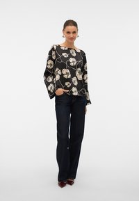 Mujer con blusa de manga larga negra con flores, vaqueros oscuros acampanados y zapatos puntiagudos burdeos, de pie con una mano en el bolsillo.