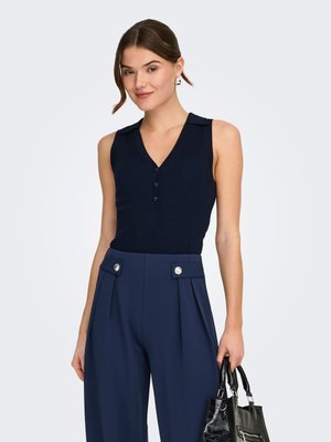 Frau trägt ein ärmelloses marineblaues Oberteil und hochtaillierte marineblaue Hosen mit aufgesetzten Taschen und Knöpfen, hält eine schwarze, strukturierte Handtasche.