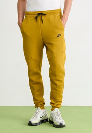 Yellow Nike sweatpants with a drawstring waist, featuring a small logo and panel design. Worn with silver Nike sneakers on a green floor.