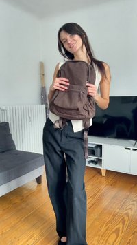 Brown fabric backpack with a zippered front pocket, adjustable straps, and a soft texture. Worn by a person in a light top and dark pants.