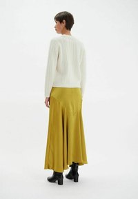 Un pull en maille blanche associé à une longue jupe fluide en satin jaune moutarde, porté avec des bottines à talons noires.