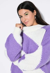 Flauschiger Strickpullover mit einem Rautenmuster in Lila und Weiß, hoher Kragen und oversized Ärmel. Die weiche Textur ist ausgeprägt.