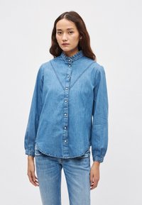 Denimskjorte, lyseblå, med lange ermer, rysjekrage, dekorativ søm, knappestolpe foran, avrundet kant, med små knapper.