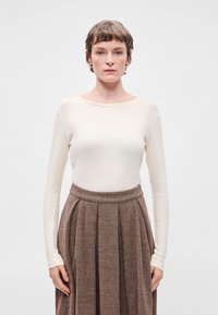 Tejfehér bordázott, hosszú ujjú felső, ami egy barna halszálkás mintázatú szoknyával van párosítva, amely pliszírozott és szűk derékkal rendelkezik. Minimalista dizájn.