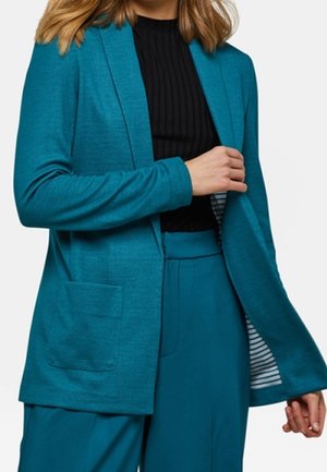 Vrouw met een blauwgroene blazer en bijpassende broek over een zwart geribd topje, houdt de blazer open om de gestreepte voering binnenin te tonen.