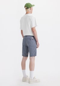 Shorts de mezclilla gris con un corte relajado, bolsillos traseros y un parche con el logo, combinados con una camiseta blanca y una gorra verde. Incluidos zapatos de color claro.