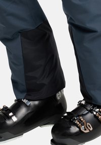 Skihosen mit einem blauen oberen Teil und einem strukturierten schwarzen unteren Teil. Aus wasserdichtem Material gefertigt und mit verstärkten Säumen. Schwarze Skischuhe mit silbernen und braunen Beschlägen.