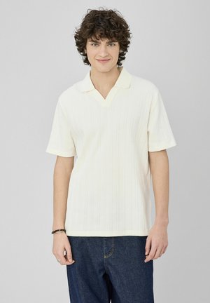 Polo à manches courtes jaune pâle avec un col en V, motif côtelé texturé, associé à un pantalon en denim foncé. Tissu doux et coupe décontractée.