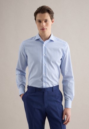 Junger Mann trägt ein hellblau gestreiftes Hemd, das in dunkelblaue Anzughosen gesteckt ist, und steht mit einer Hand in der Tasche.