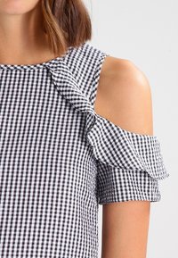 Czarna i biała bluzka w kratkę gingham z wycięciami na ramionach, falbanami na rękawach i dopasowanym dekoltem. Materiał z mieszanki bawełny.