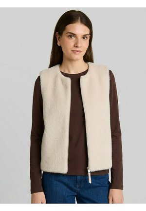 Jeune femme aux longs cheveux bruns porte un gilet duveteux couleur crème par-dessus une chemise marron à manches longues et un jean bleu sur un fond uni.