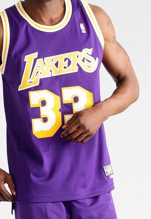 Man draagt een paarse basketbaltrui en korte broek van de Los Angeles Lakers met nummer 33, en past de trui aan met zijn rechterhand.