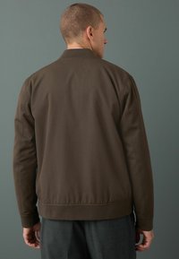 Chaqueta bomber marrón con un cuello y puños acanalados, con un diseño minimalista y una textura suave. Vista trasera con dobladillo elástico.