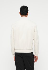 Jaqueta bomber em bege claro, com colarinho e punhos canelados, cintura elástica, tecido suave e design minimalista nas costas.