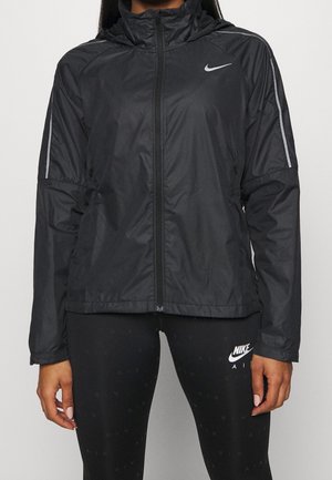 Osoba ubrana w czarną kurtkę Nike zapinaną na zamek z odblaskowymi paskami na rękawach oraz czarne legginsy Nike z białymi logo, stojąca na jednolitym tle.