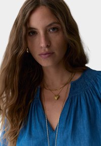 Collana a pendente a forma di cuore dorato sovrapposta su una persona che indossa una maglietta in denim blu con scollatura arricciata e tessuto strutturato, sfocata.