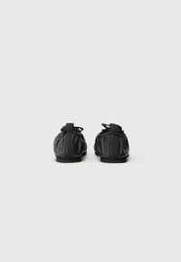 Schwarze, weiche Lederschuhe mit abgerundetem Rücken, elastischem Design und dezentem Branding an der Ferse. Flache Sohle mit minimaler Textur.