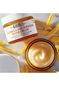 Kiehl's Calendula Serum-Infused Water Cream i en klar krukke med et gult låg, omgivet af lyse gule kalendulablade på en glat overflade.