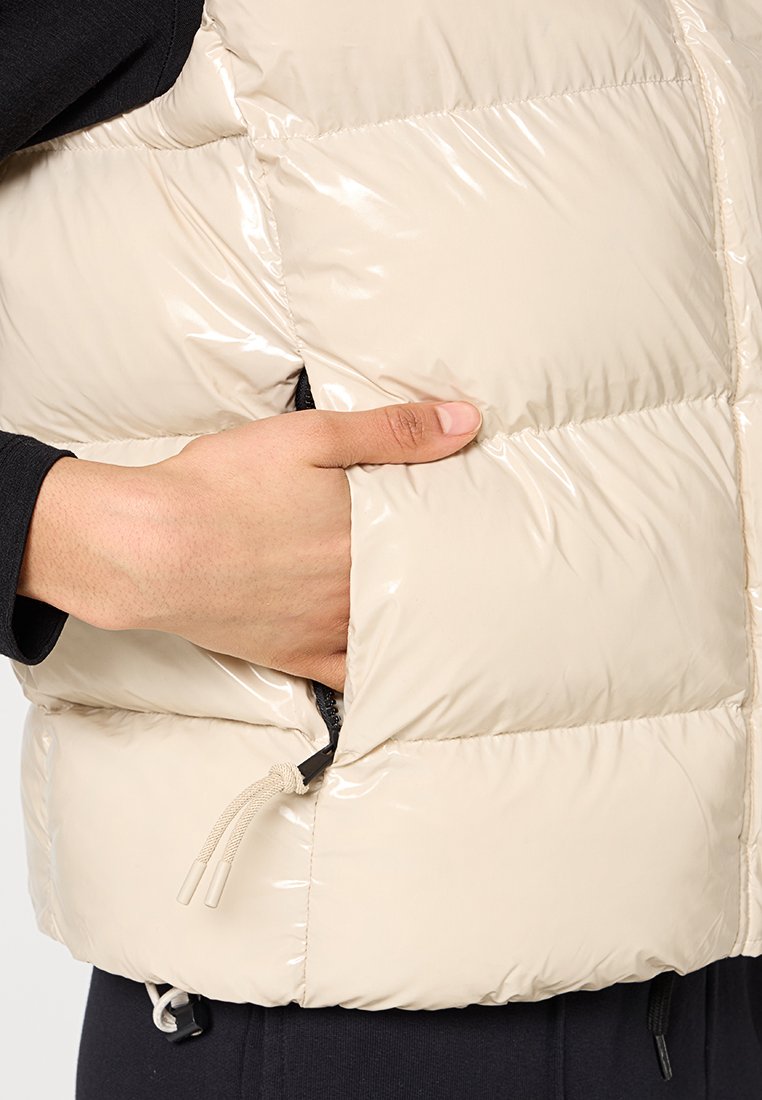 Hand in einer beigen Puffer-Weste mit sichtbarem Reißverschluss und Kordelzug, die über einem langärmeligen schwarzen Oberteil getragen wird.