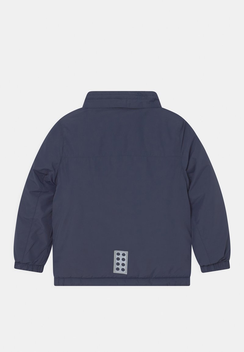 Dunkelblau gesteppte Jacke mit spitzem Kragen, elastischen Bündchen und einem Rückendesign, das einen grauen Patch mit runden Ausschnitten zeigt.