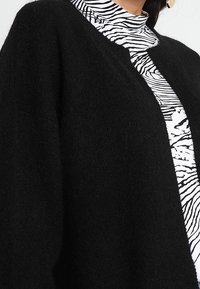 Czarny dzianinowy kardigan z otwartym przodem, o miękkiej fakturze. Pod nim znajduje się top z wysokim kołnierzem w czarno-białym wzorze w zebrę.