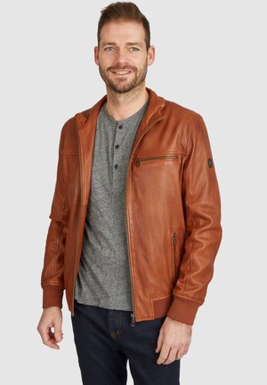 Chaqueta de cuero marrón con cremallera frontal, puños acanalados y un bolsillo en el pecho. Llevada sobre una camiseta tipo henley gris, con un ajuste casual.