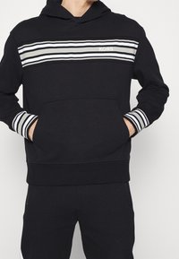 La felpa nera presenta una tasca frontale, dettagli a righe in grigio e bianco e un logo sul petto in un colore a contrasto. Tessuto morbido.