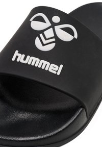 Svart slingback sandal i texturerat gummi med en bred rem som har en vit logotyp och designdetaljer. Platt sula för komfort.