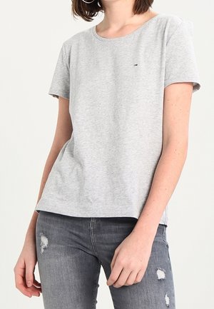 Vrouw die een lichtgrijs T-shirt met korte mouwen en verweerde donkergrijze jeans draagt, staand tegen een effen achtergrond.
