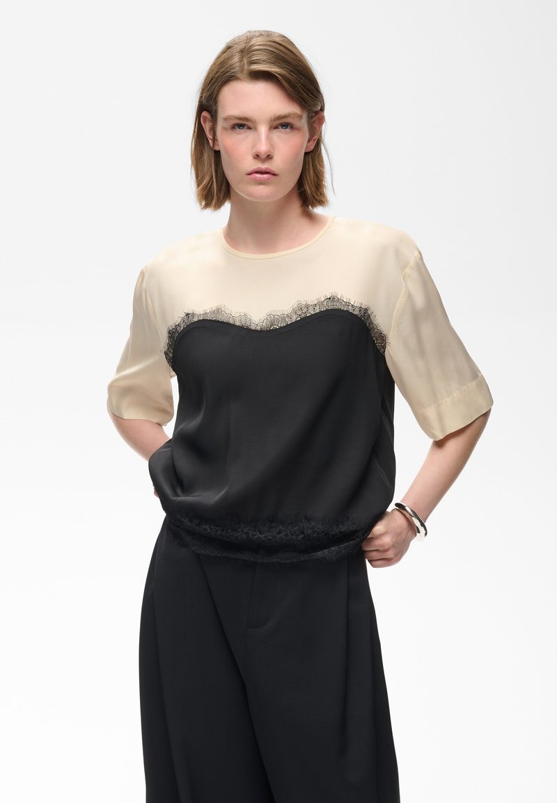 Jeune personne aux cheveux courts châtain clair porte un haut beige et noir avec dentelle et un pantalon noir, debout avec une main sur la hanche.