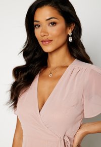 Vestito avvolgente rosa blush con maniche corte, tessuto texturizzato, scollatura a V e un fiocco in vita. Accessoriato con orecchini statement e una collana.