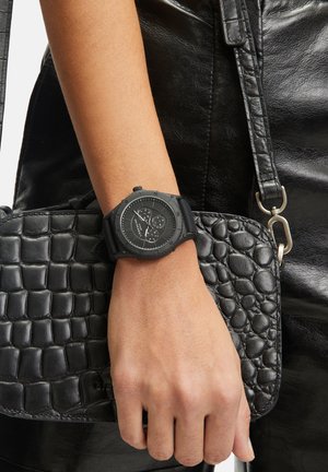 Montre noire avec cadran texturé et bracelet en silicone, à côté d'un sac noir à motif crocodile avec des accents en cuir lisse.