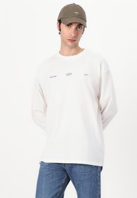 White long-sleeve t-shirt with blue text and graphic designs, paired with blue jeans and a green cap featuring white embroidery.